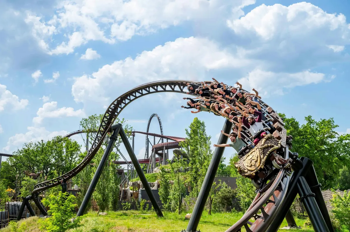 Des montagnes russes avec des passagers sont en mouvement dans une boucle, dans un parc d'attractions, au milieu de la verdure et sous un ciel partiellement nuageux.
