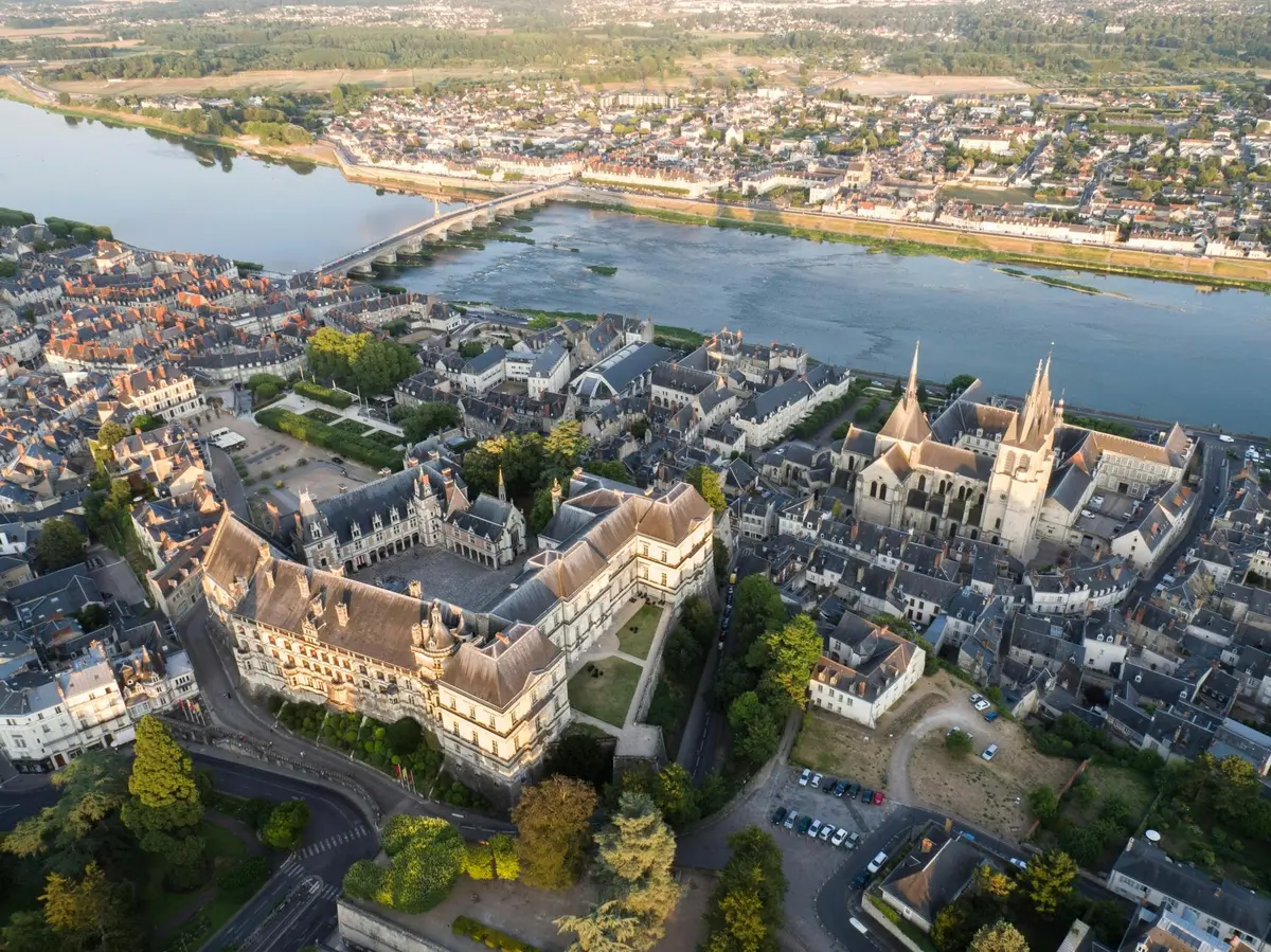 Vue aérienne d'une ville européenne historique avec une rivière, une cathédrale et des bâtiments ressemblant à des châteaux, entourés d'arbres et de maisons.