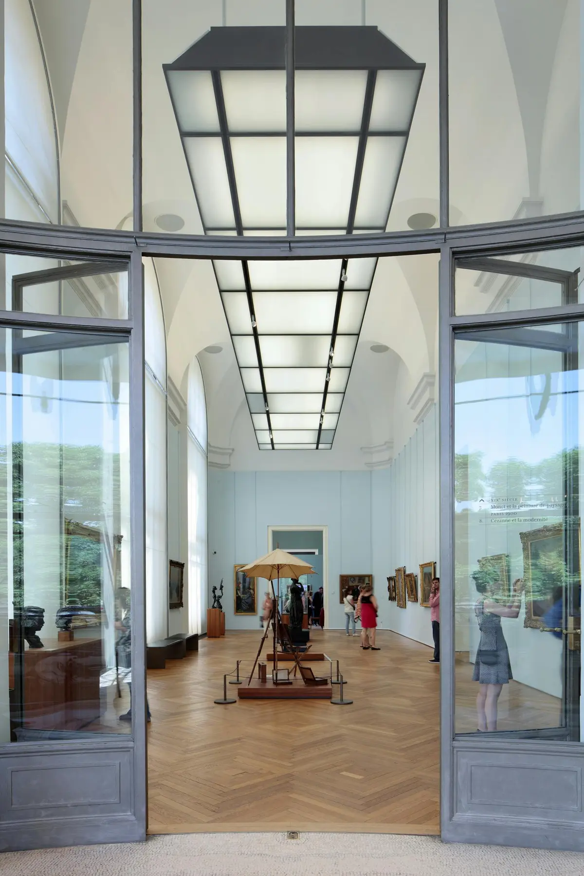 Museum gallery with high ceilings, glass walls, and visitors viewing paintings and sculptures. Central installation under an umbrella.