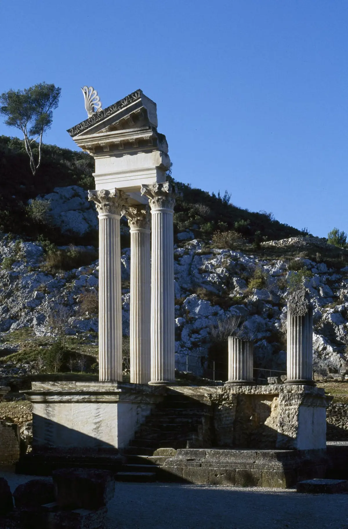 Ruines anciennes avec de hautes colonnes blanches et une structure partielle sur un terrain rocheux sous un ciel bleu clair. Des arbres se trouvent à l'arrière-plan.