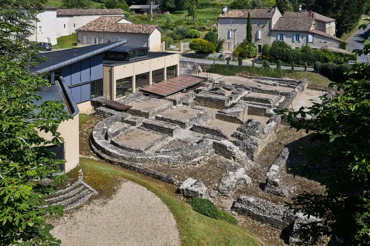 Ruines archéologiques avec des fondations en pierre, entourées de bâtiments modernes et traditionnels avec des toits en tuiles, avec de la verdure en arrière-plan.