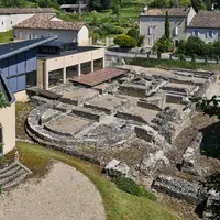 Ruines archéologiques avec des fondations en pierre, entourées de bâtiments modernes et traditionnels avec des toits en tuiles, avec de la verdure en arrière-plan. &copy; © Damien Lachas / Centre des monuments nationaux