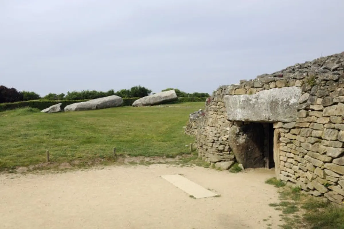 Structure en pierre avec, en arrière-plan, un champ herbeux contenant de grandes formations rocheuses, sous un ciel nuageux.