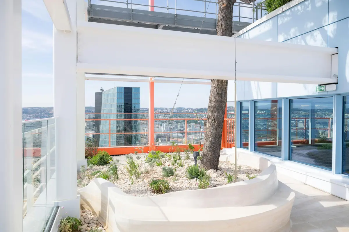 Un jardin sur le toit avec des jardinières blanches, un grand arbre et des vues sur le paysage urbain avec un immeuble de grande hauteur bleu et des garde-corps orange.