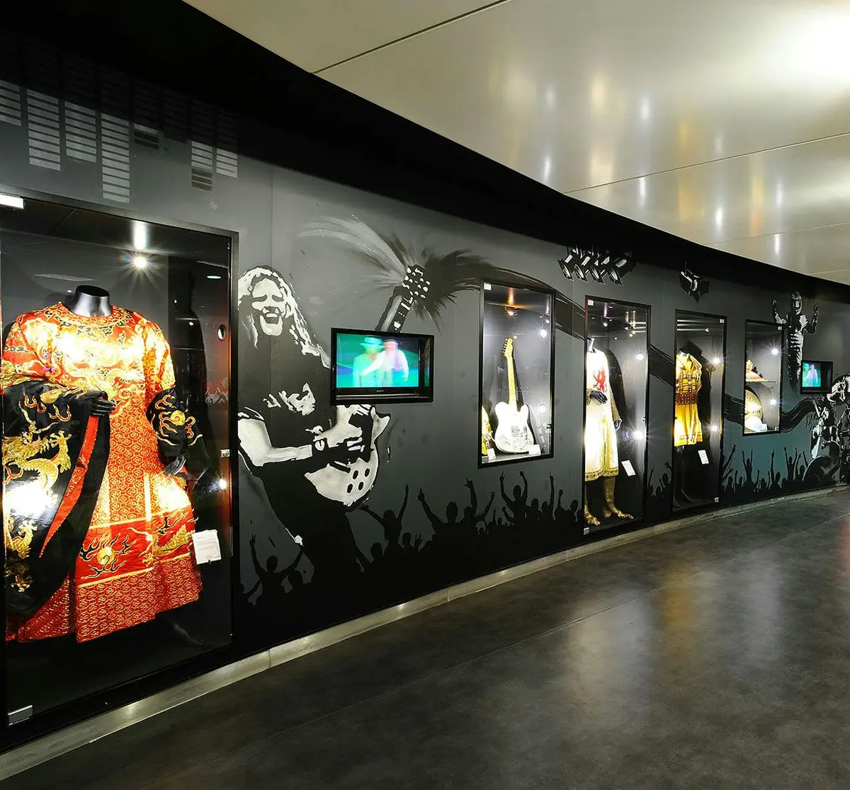 Un mur d'exposition de musée présentant des souvenirs de rock, avec des costumes, des guitares et un écran vidéo.