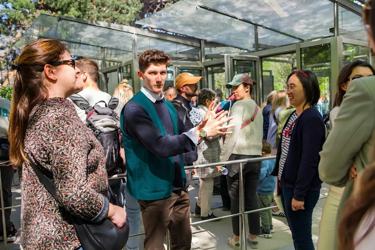 Des personnes sont alignées à l'extérieur, avec un homme qui fait des gestes au premier plan. On aperçoit des structures en verre et des arbres.
