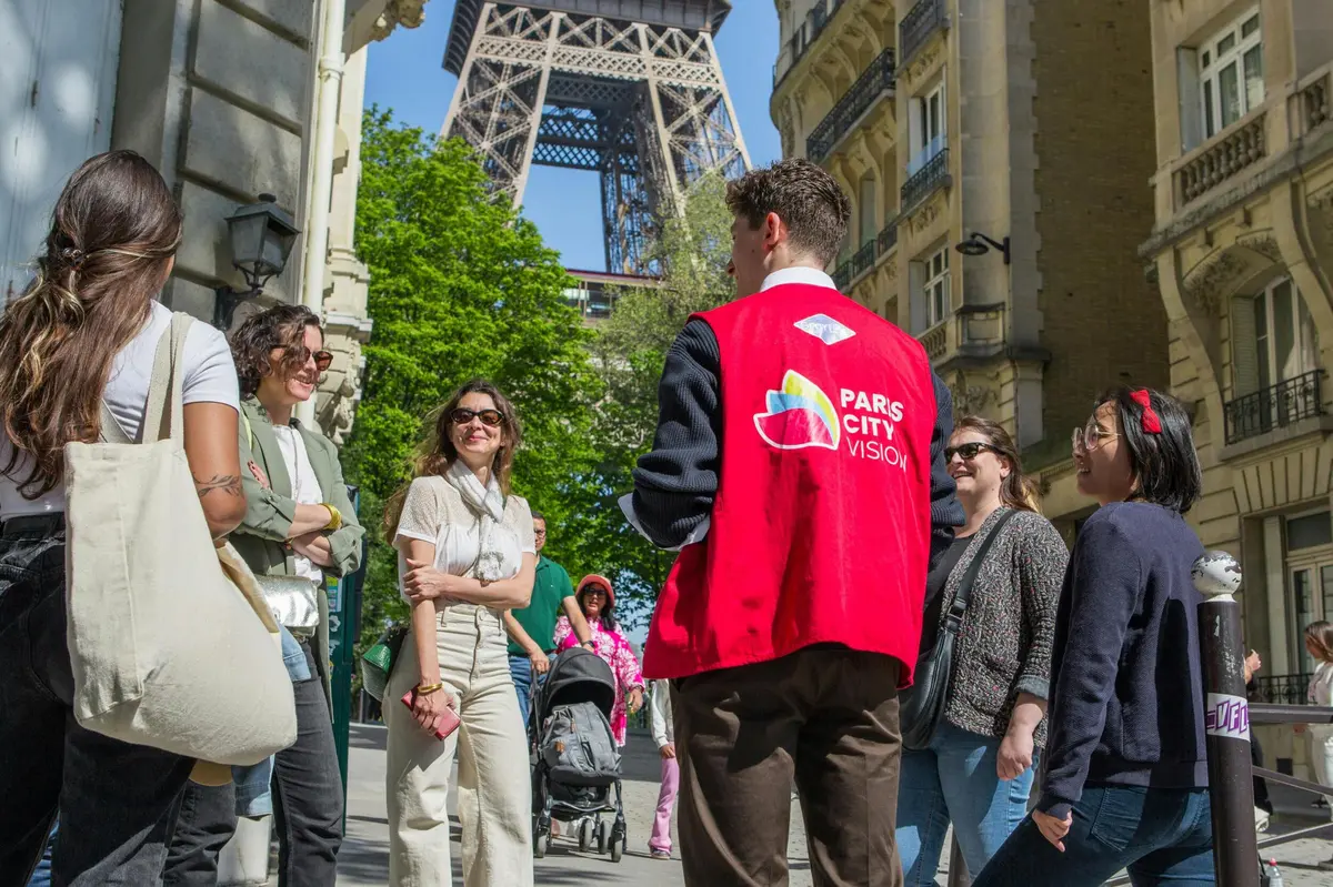 Un guide portant un gilet rouge étiqueté Paris City Vision s'adressant à un groupe de touristes près de la Tour Eiffel lors d'une journée ensoleillée.