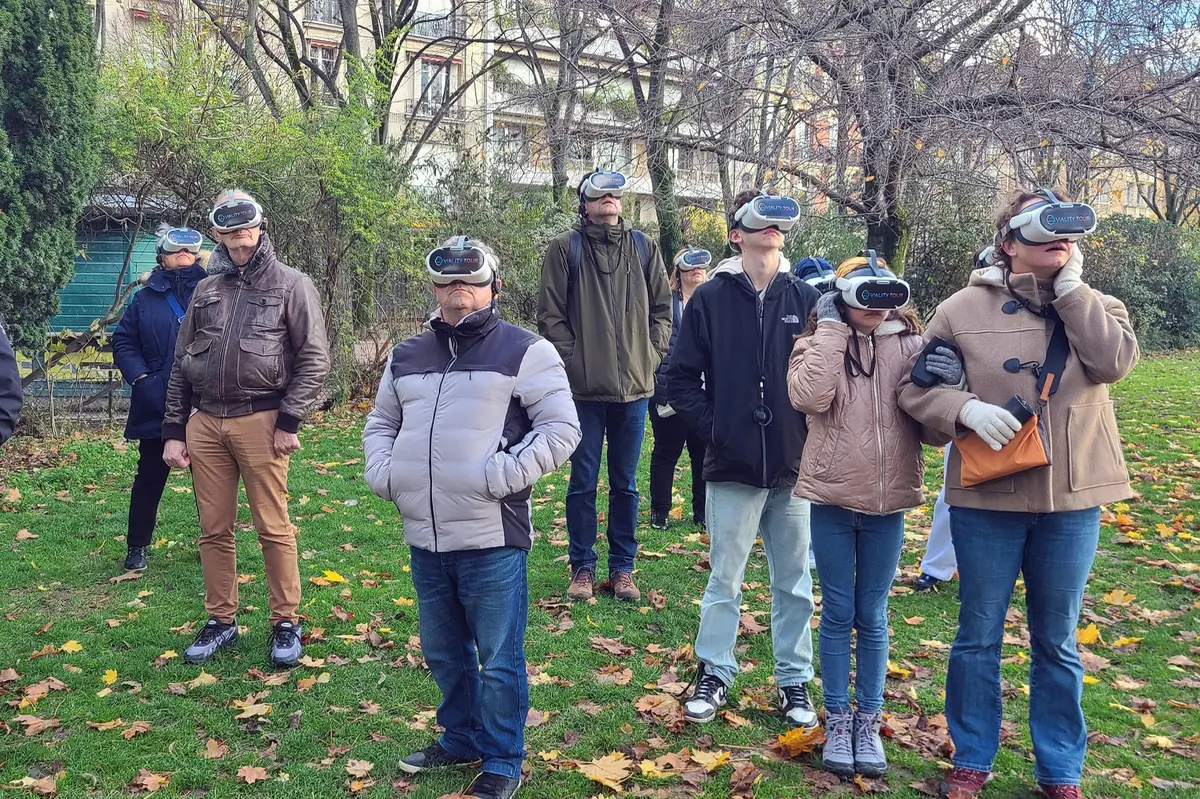 A group of people outdoors wearing VR headsets, standing on a grassy area surrounded by trees and fallen leaves.