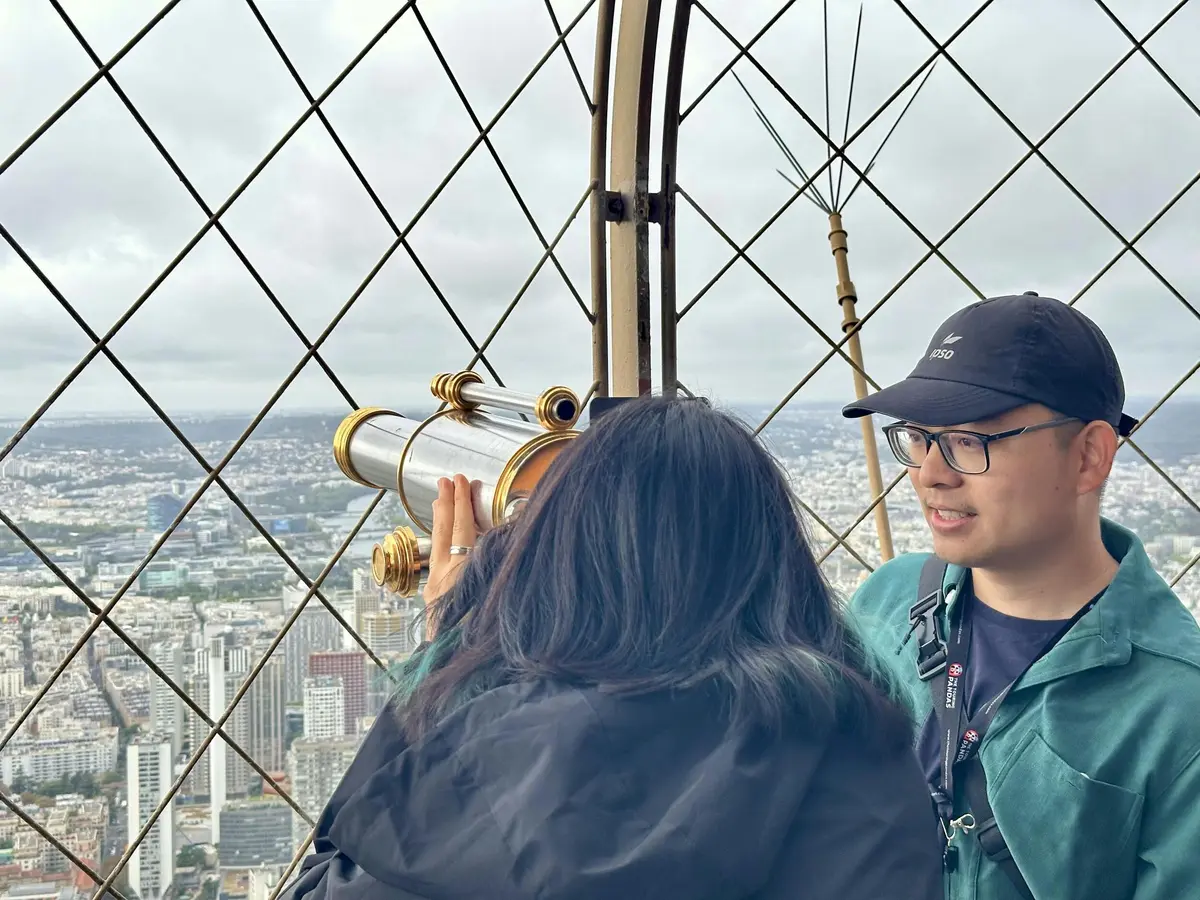 Deux personnes sur un pont d'observation avec des barres métalliques et un télescope, surplombant un paysage urbain sous un ciel nuageux.