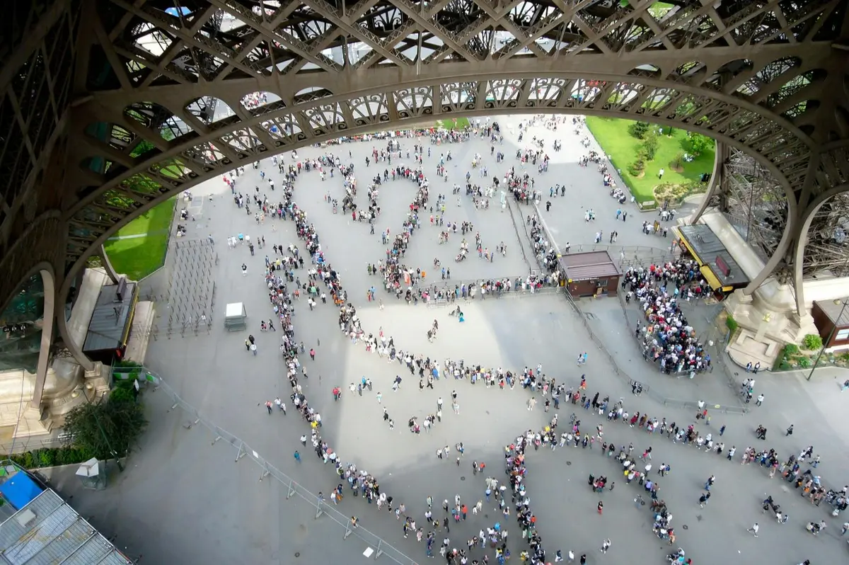 Vue aérienne montrant de longues files d'attente serpentant au pied de la Tour Eiffel, avec de la verdure et une place en arrière-plan.