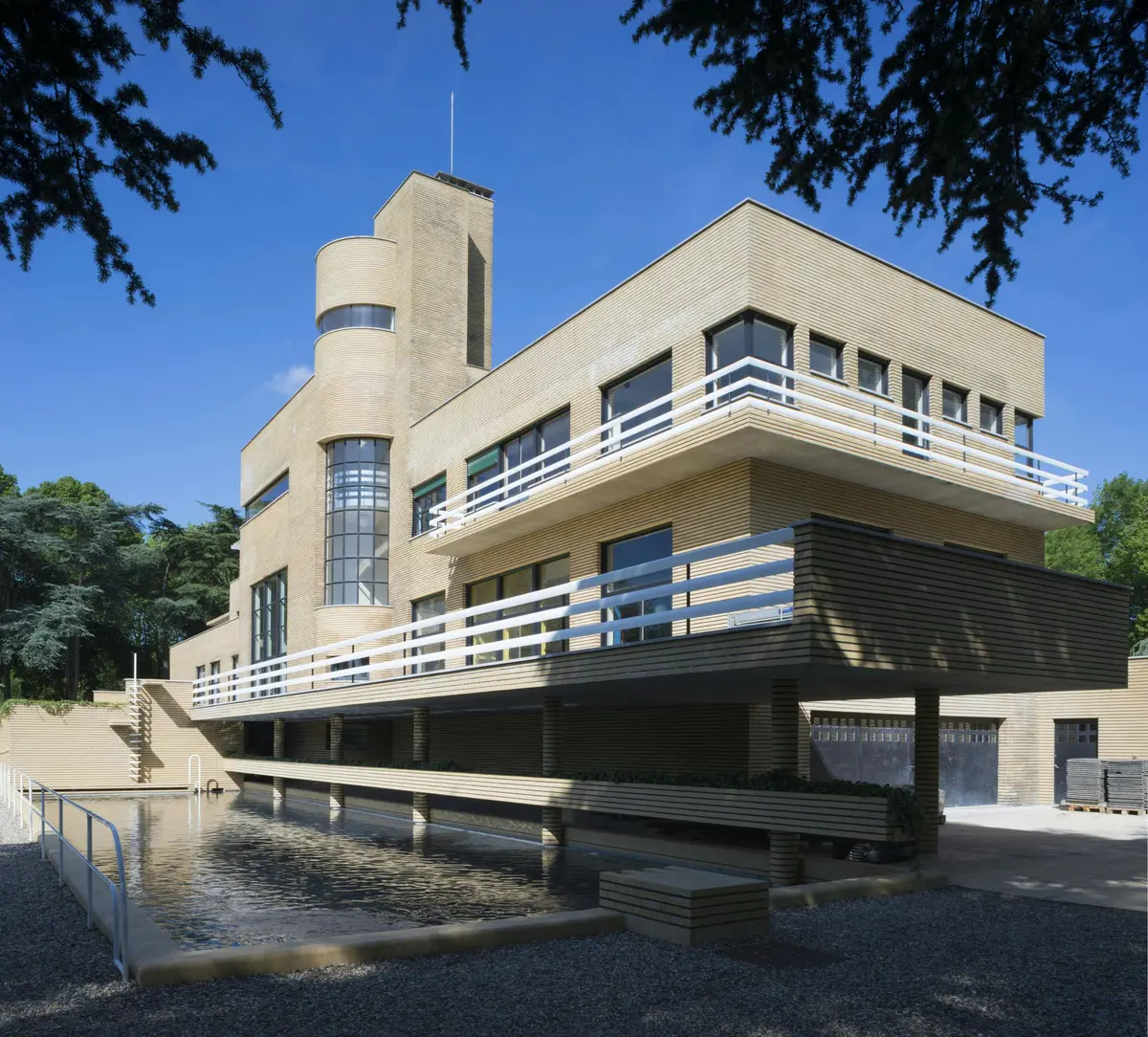 Bâtiment moderne de plusieurs étages avec un extérieur en briques, de grandes fenêtres et des balcons. Piscine réfléchissante au premier plan.