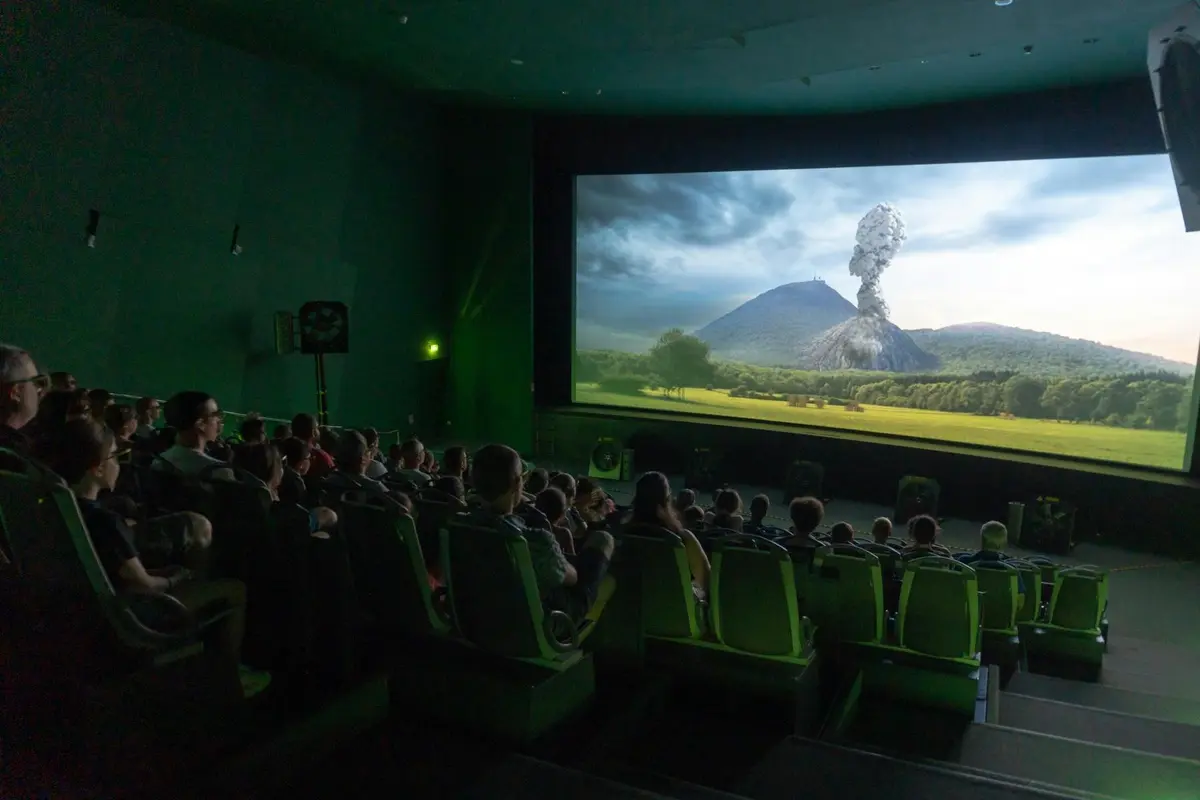 Des personnes assises dans une salle obscure regardent une scène de film mettant en scène une éruption volcanique dans un paysage.