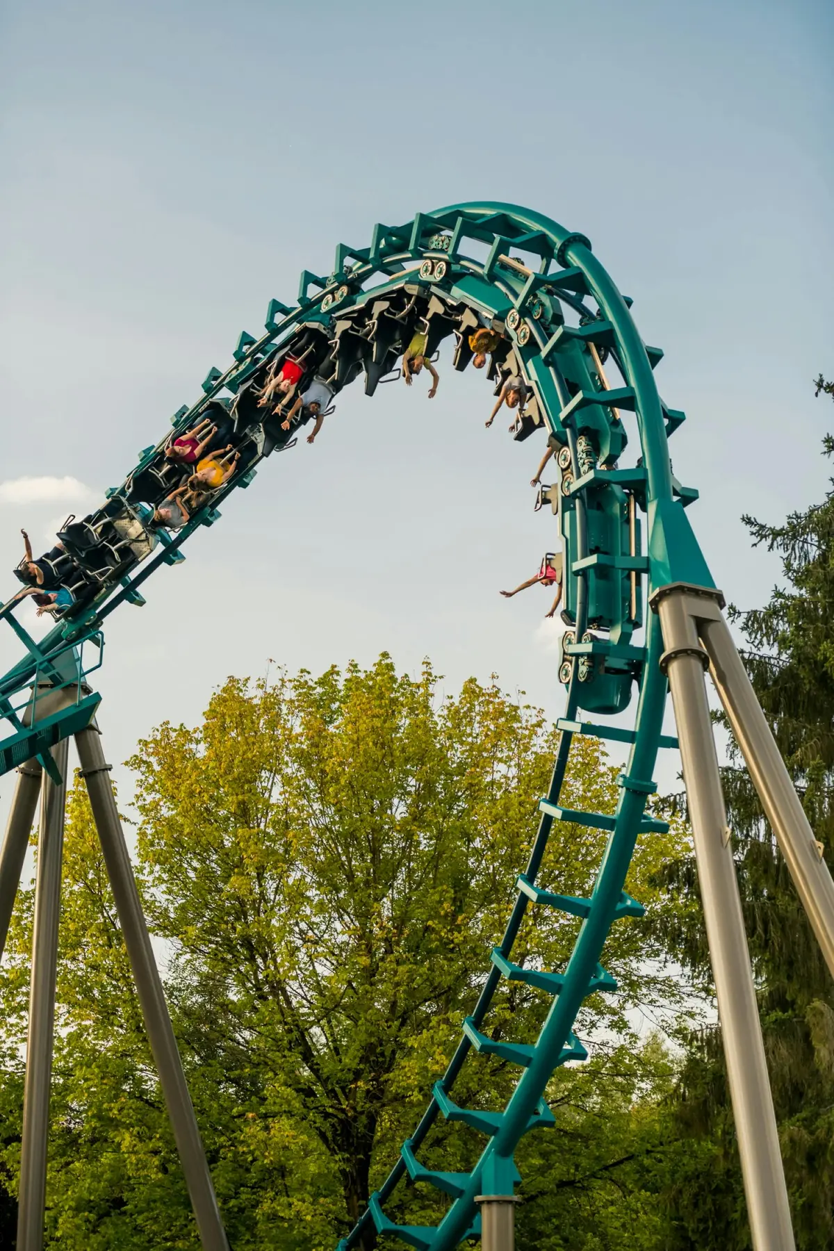 Des personnes montent sur des montagnes russes en boucle dont la piste est de couleur sarcelle, sur fond d'arbres verts et de ciel clair.