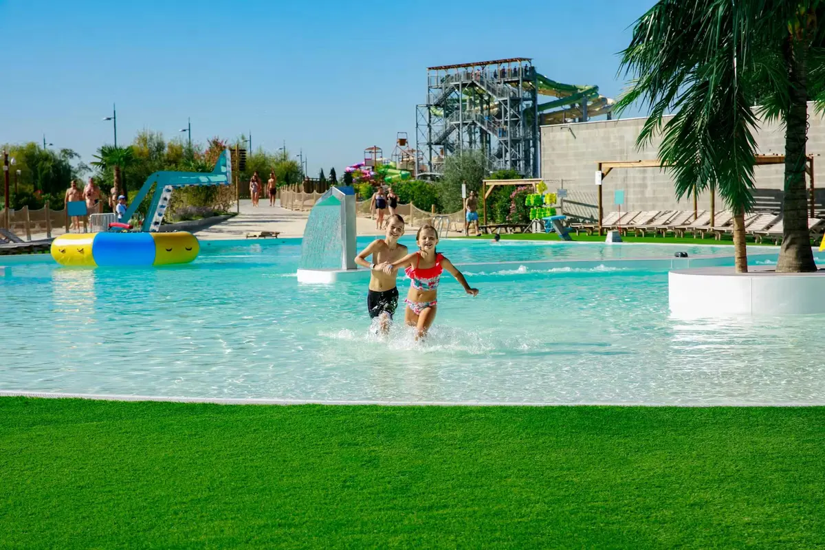 Deux enfants jouent dans une pataugeoire d'un parc aquatique, au milieu d'une végétation luxuriante, de toboggans et sous un ciel bleu.