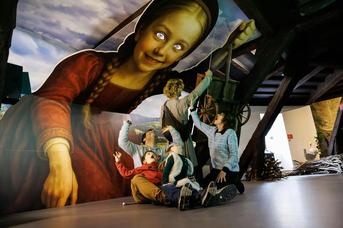 Une famille interagit avec une grande fresque surréaliste représentant une jeune fille aux yeux brillants, sur laquelle figurent un rouet et des éléments en bois.