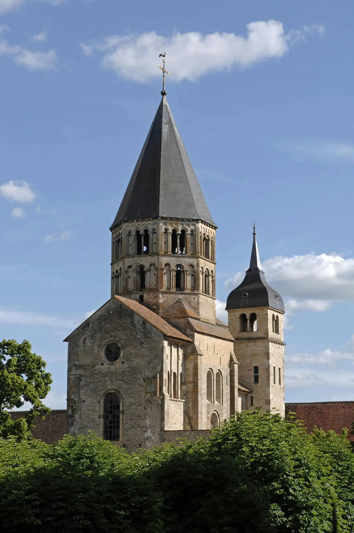 Église en pierre avec une tour centrale haute et pointue et un clocher rond plus petit, sur fond de ciel bleu avec quelques nuages.