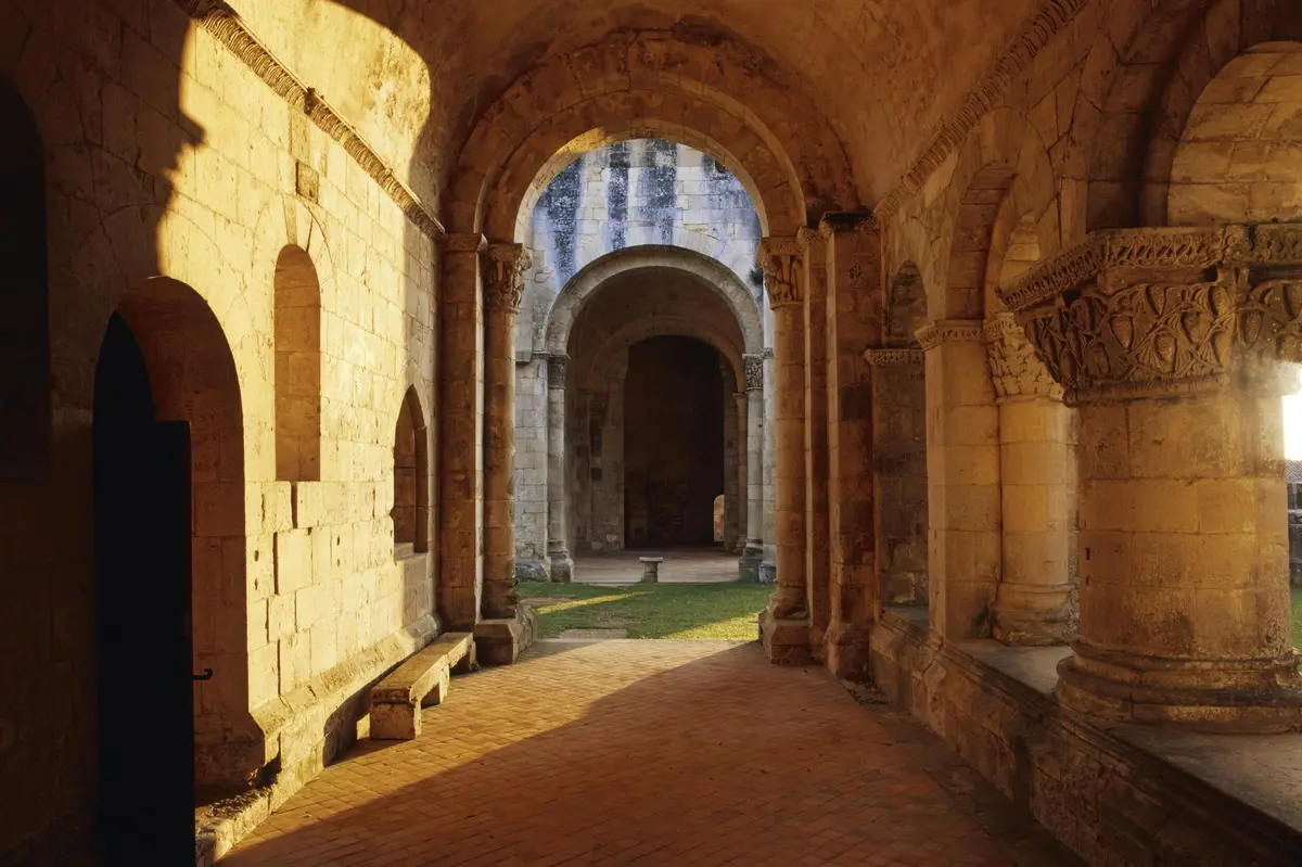 Arcades en pierre éclairées par le soleil dans un cloître médiéval avec une allée en briques et une cour herbeuse.