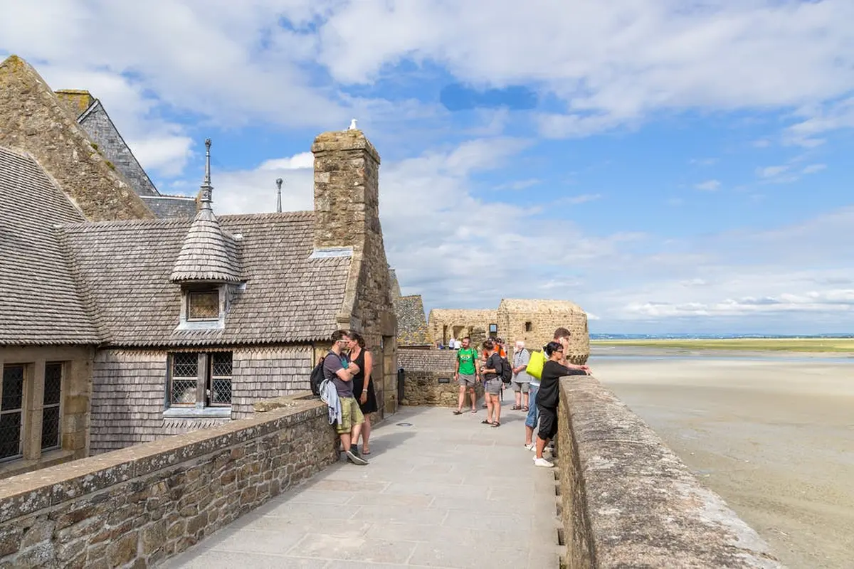 Personnes debout et assises sur un sentier en pierre surplombant une vue panoramique près de bâtiments historiques en pierre, par une journée claire et ensoleillée.