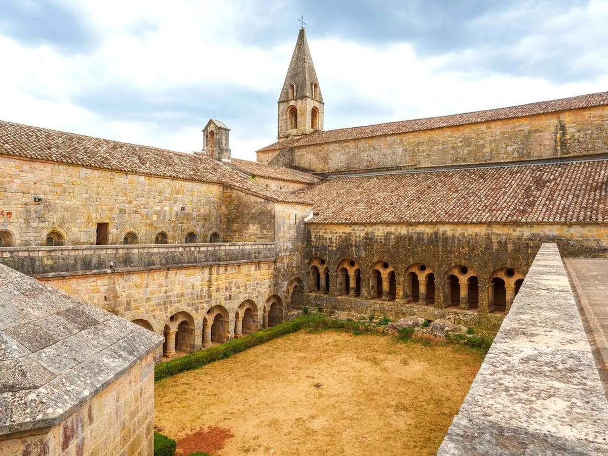 Ancienne cour de monastère en pierre avec des allées voûtées, une zone centrale d'herbe sèche et une haute flèche sous un ciel nuageux.