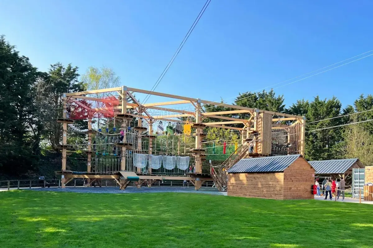 Parcours d'aventure en plein air avec des plateformes en bois, des cordes, des filets, des tyroliennes et une petite cabane en bois ; entouré d'herbe et d'arbres.