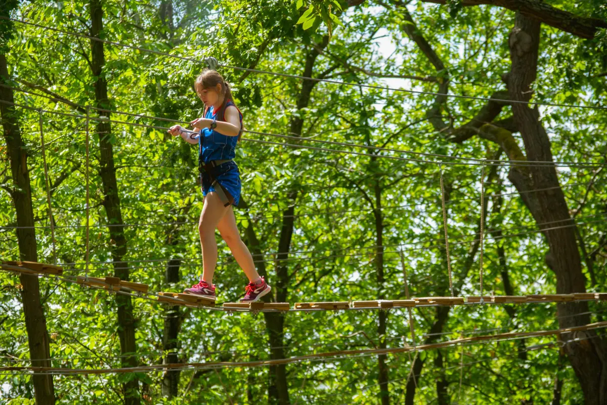 Une jeune fille équipée d'un harnais et d'un casque marche prudemment sur un pont de corde entre les arbres d'un parc d'aventure en forêt.