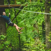 Personne souriante faisant de la tyrolienne dans une forêt luxuriante, portant un harnais et un équipement de sécurité. Une plate-forme en bois surélevée est visible à l'arrière-plan. &copy; AccroCamp