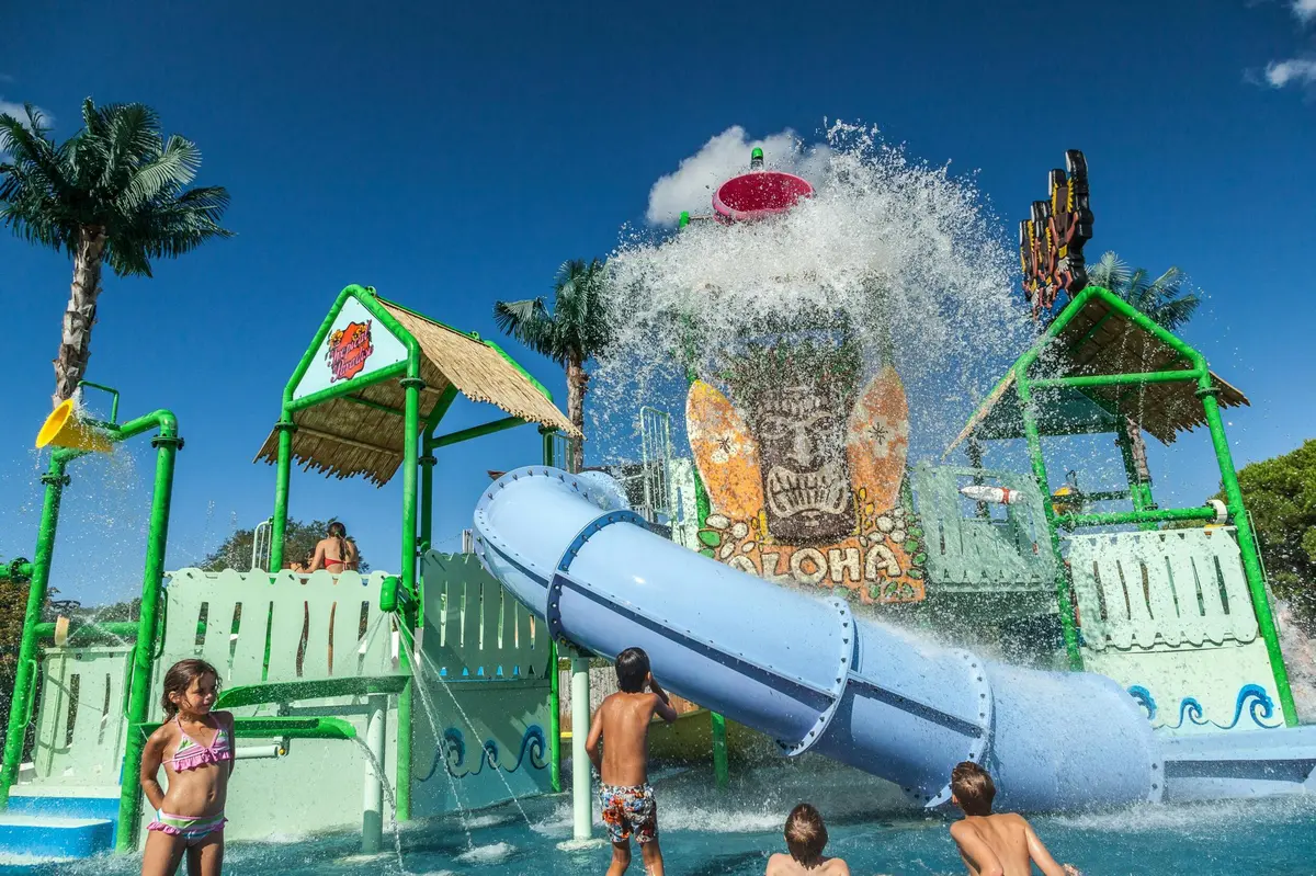 Des enfants s'amusent dans un parc aquatique haut en couleur, avec des toboggans, des palmiers et un grand seau qui déverse de l'eau, sous un ciel dégagé.