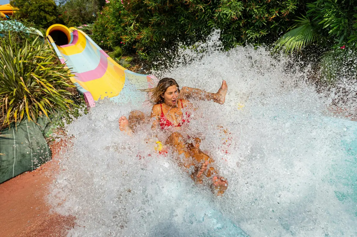 Une personne sur un toboggan aquatique coloré s'éclaboussant dans la piscine en bas, entourée de verdure.