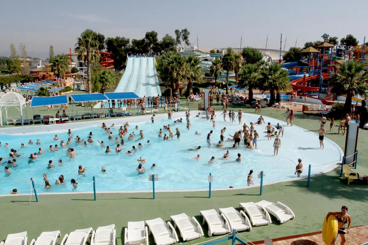 Parc aquatique bondé avec des gens qui nagent dans une grande piscine, de grands toboggans, des palmiers et de nombreuses chaises longues.