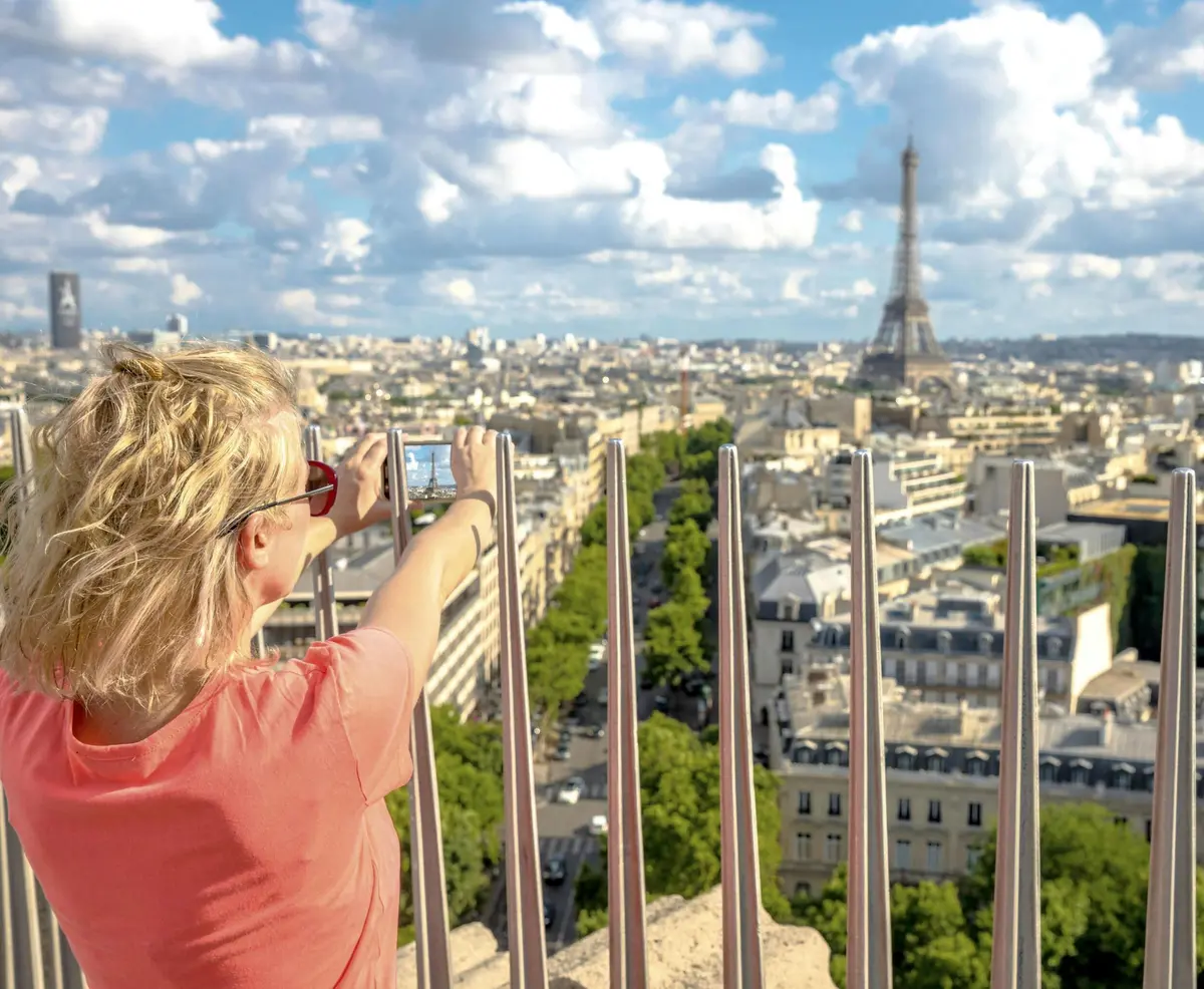 Une femme portant des lunettes de soleil prend une photo de la Tour Eiffel et de la ligne d'horizon de Paris par une journée ensoleillée, vue à travers une balustrade.