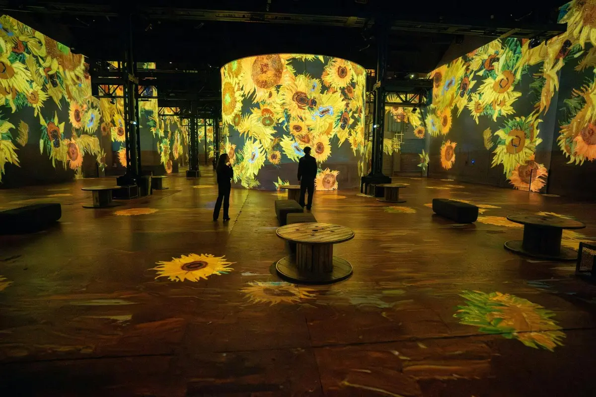 Une installation artistique immersive mettant en scène de grands tournesols numériques projetés sur les murs et le sol, devant un public de spectateurs.