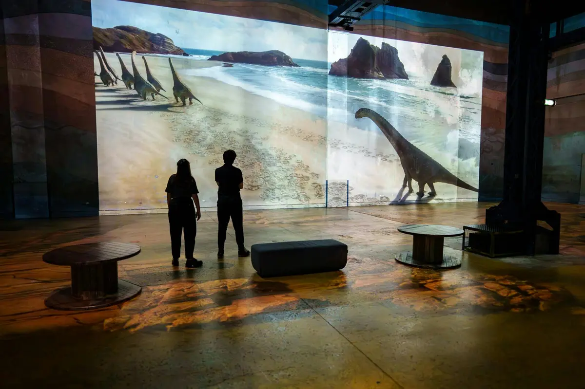 Deux personnes se trouvent dans une galerie et observent une grande projection de dinosaures marchant sur une plage, avec des sièges et des décors autour.