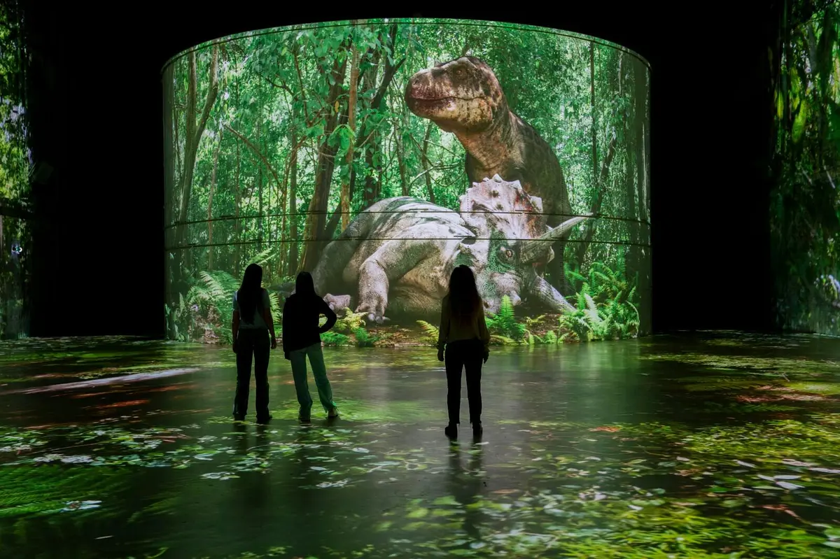 Trois personnes se tiennent devant un grand écran numérique sur lequel est projetée une scène de forêt mettant en scène deux dinosaures : un T. rex et un tricératops.