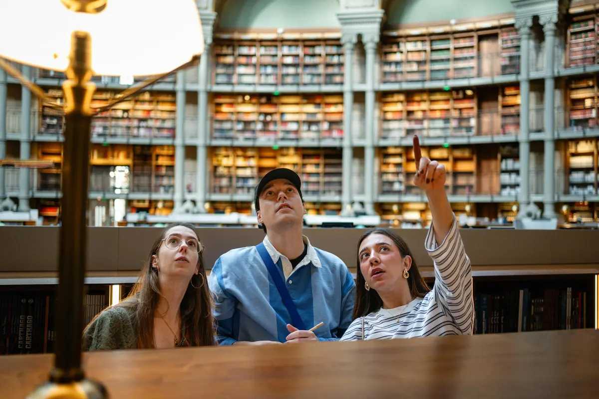 Trois personnes assises à une table dans une bibliothèque, regardant vers le haut, avec des étagères et une architecture en arc de cercle à l'arrière-plan.