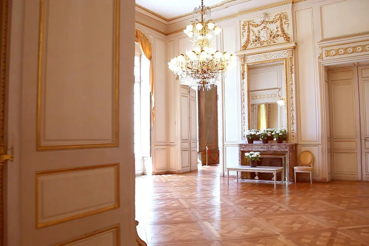 Pièce ornée d'un lustre, d'un grand miroir au-dessus d'une cheminée, de fleurs sur le manteau et de parquet. Fenêtres ensoleillées avec rideaux.