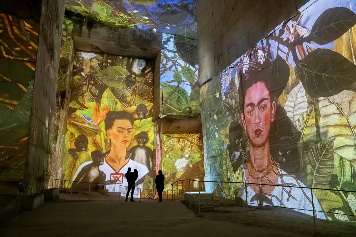 Projection d'une œuvre d'art colorée représentant un portrait entouré de feuillage sur les murs d'un grand espace intérieur avec des visiteurs.