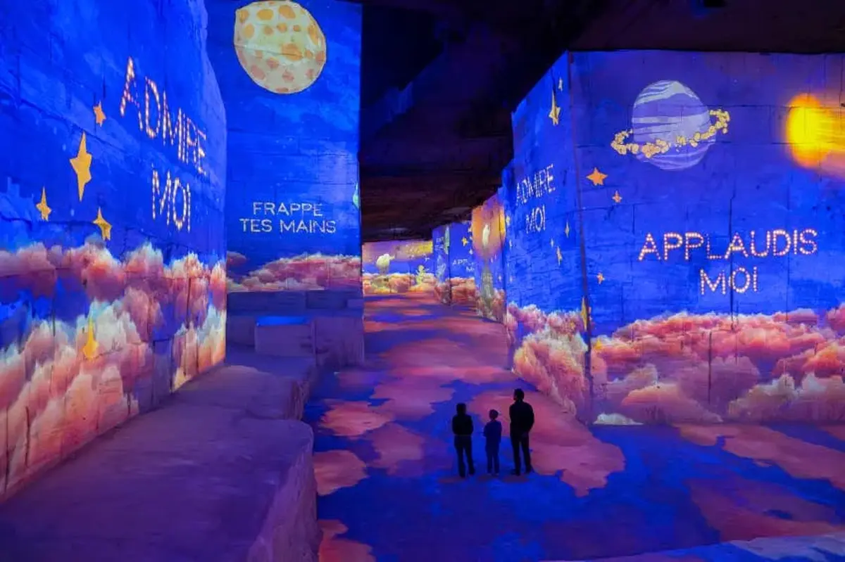 Une exposition intérieure colorée avec des projections de nuages, d'étoiles et de textes sur les murs. Les silhouettes de trois personnes sont visibles.