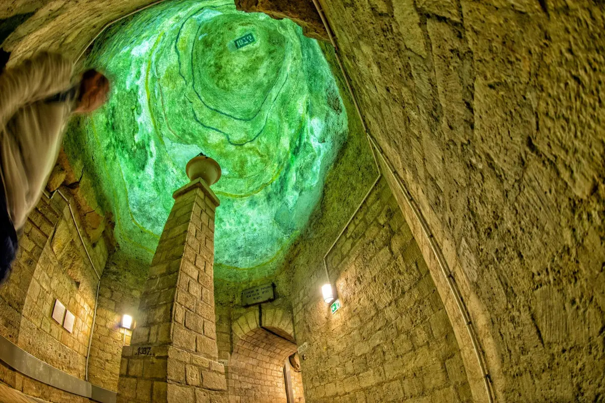 Une chambre historique en pierre avec un plafond éclairé en vert et une entrée voûtée, avec un pilier central et des lampes murales.