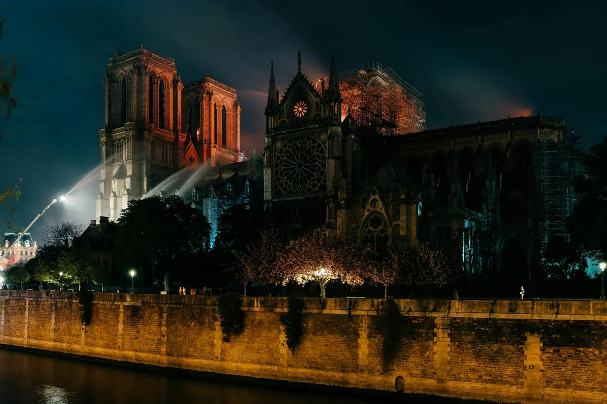 Vue nocturne de la cathédrale Notre-Dame illuminée avec, au premier plan, des échafaudages, des arbres et une berge.