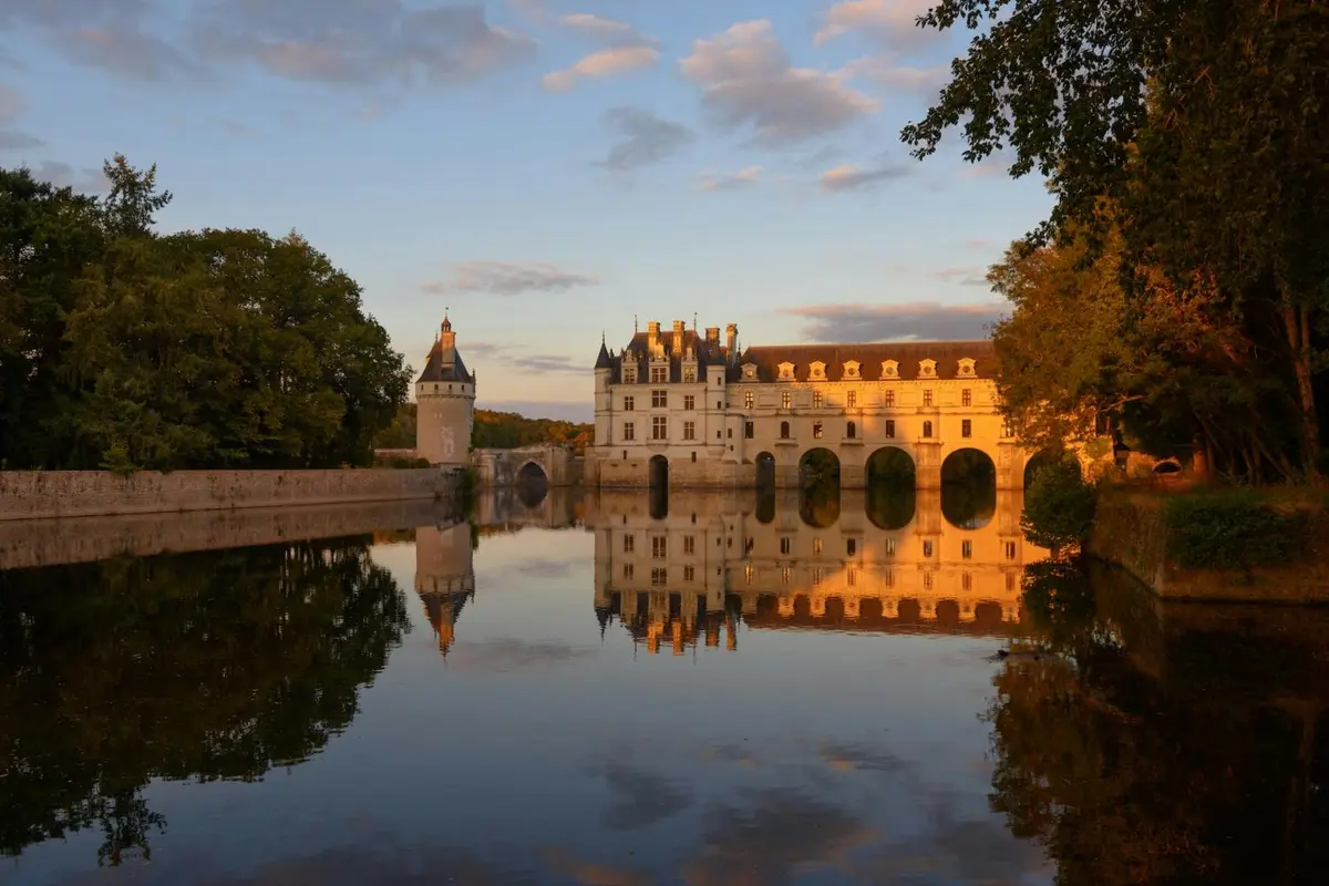 Un château avec des tours et des arches se reflétant dans une rivière calme au coucher du soleil, entouré d'arbres sous un ciel partiellement nuageux.