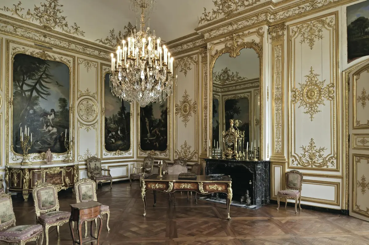 Salle ornée d'un grand lustre, de meubles anciens, de miroirs dorés et de peintures murales décoratives.