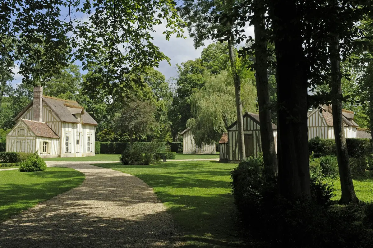 Un chemin de gravier ombragé traverse une zone herbeuse où se trouvent plusieurs chalets en bois entourés d'arbres sous un ciel partiellement nuageux.