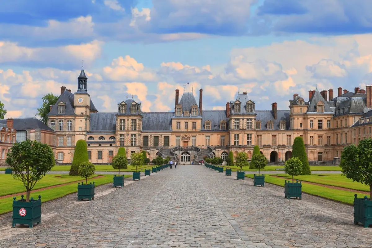Un palais symétrique, dont l'entrée est précédée d'une allée pavée, entouré de pelouses impeccablement entretenues et d'arbres, sous un ciel bleu.