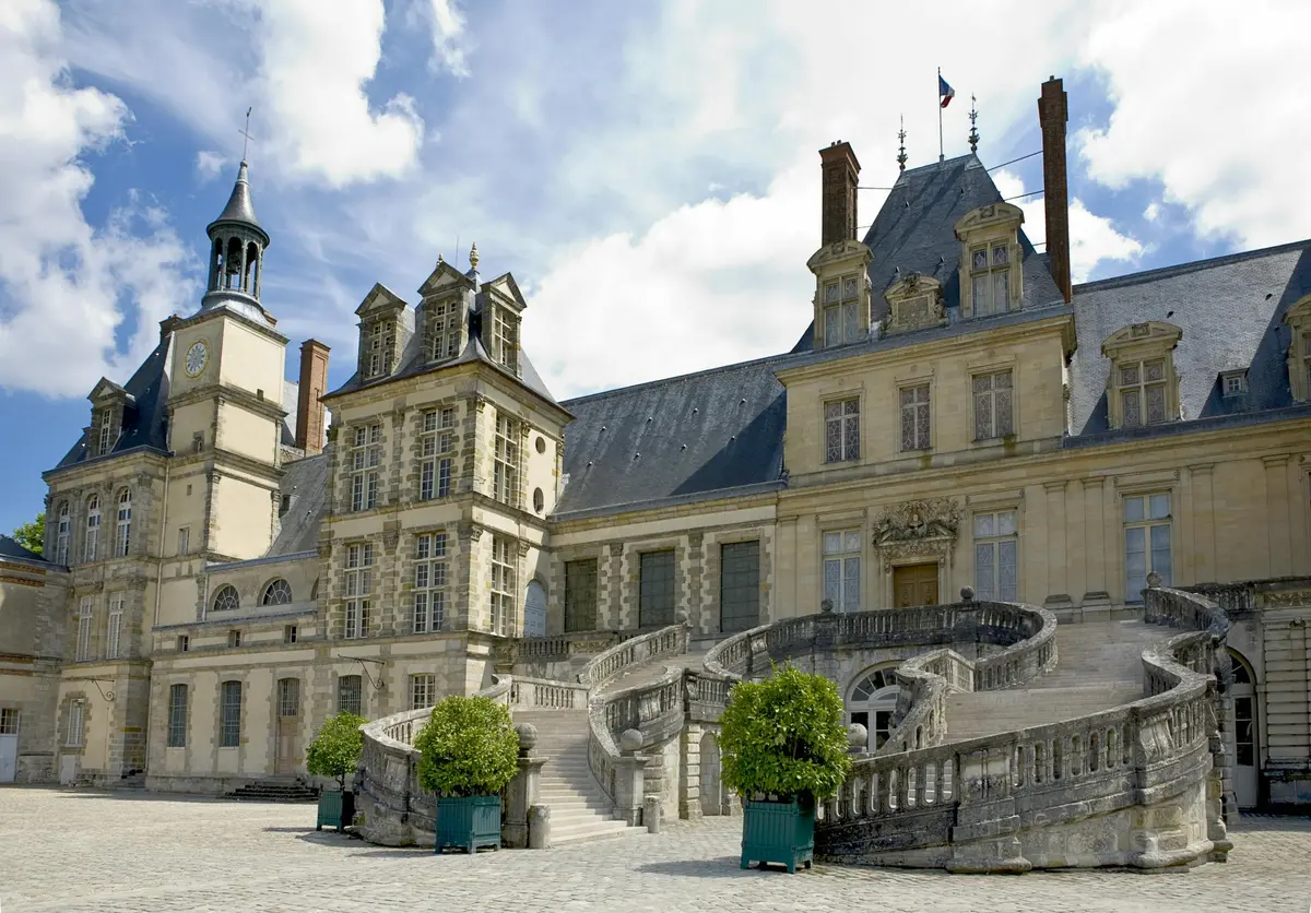 Historic chateau with ornate architecture, tall windows, a grand staircase, and manicured potted plants against a blue sky.