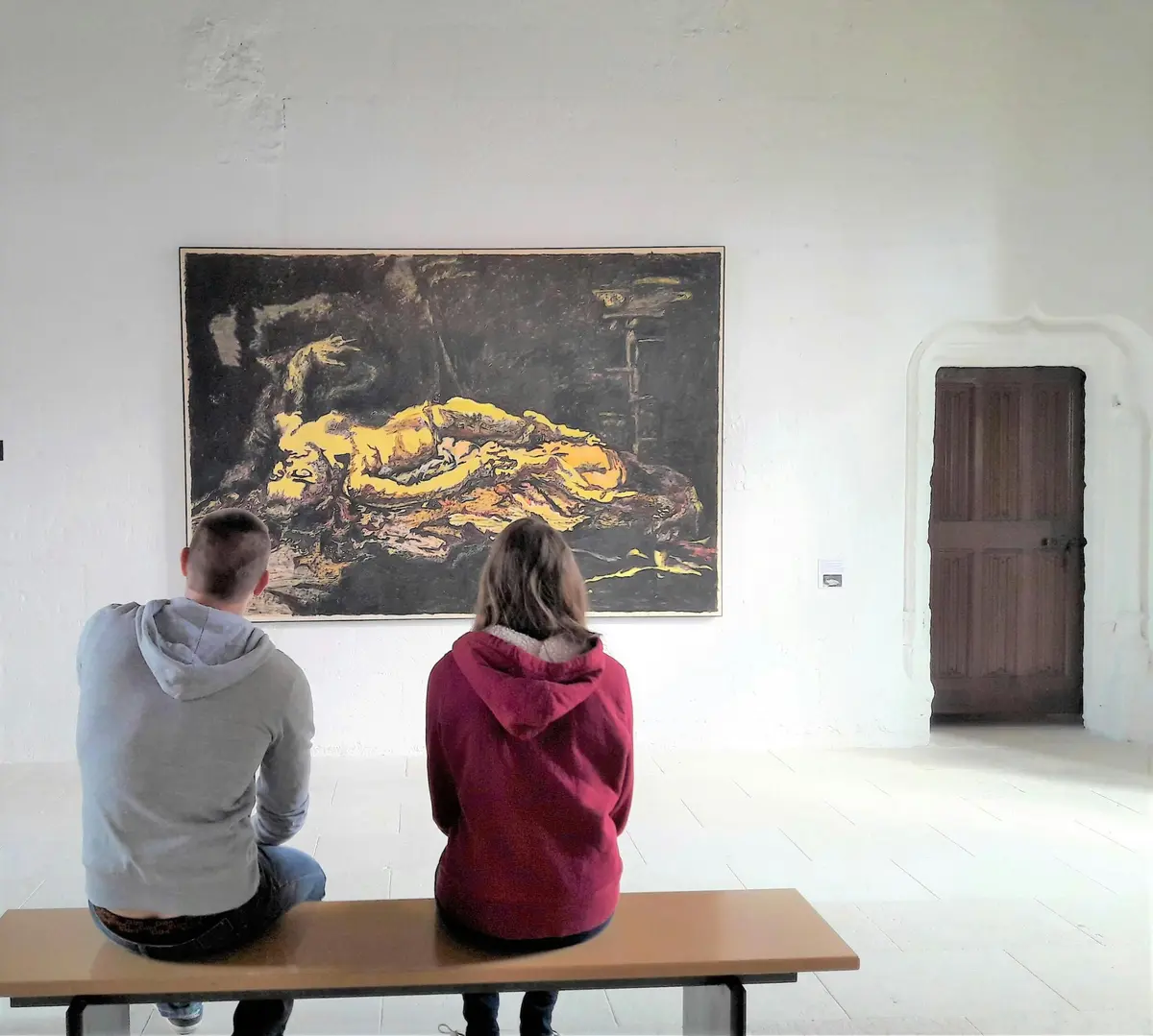 Deux personnes sont assises sur un banc et regardent un grand tableau dans une salle de musée aux murs blancs et à la porte en bois.