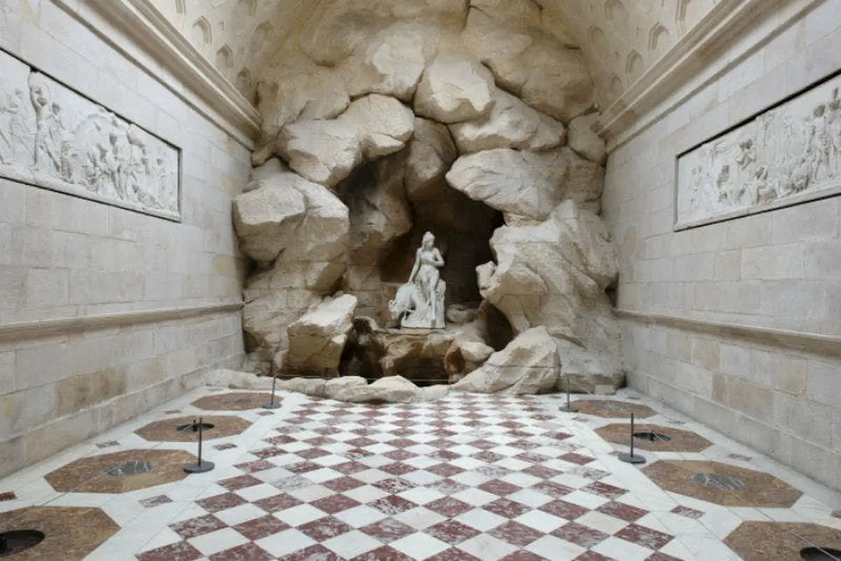 Statue d'une femme et d'un enfant dans une alcôve rocheuse au fond d'un couloir au sol en damier dans une pièce aux murs de pierre.