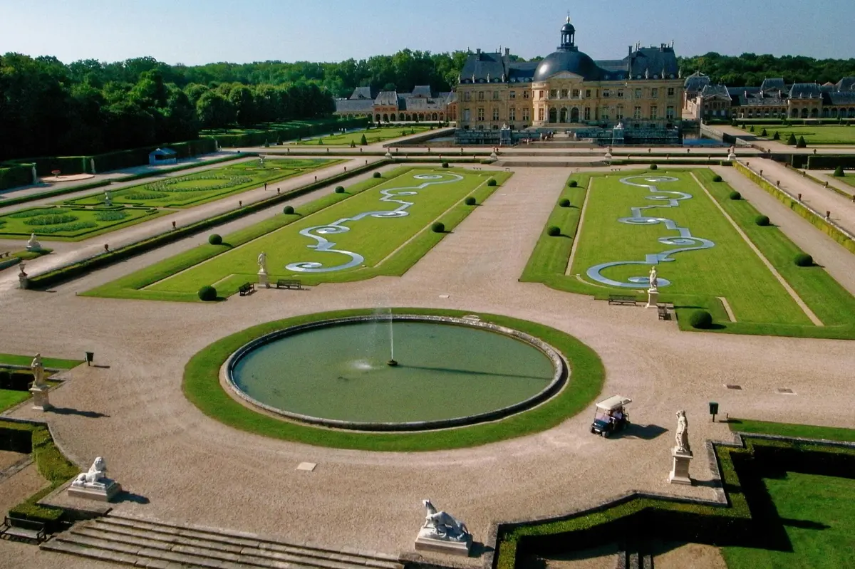 Un jardin à la française avec des pelouses symétriques, des allées décoratives et une fontaine ronde. Un grand bâtiment en forme de dôme se trouve à l'arrière-plan.