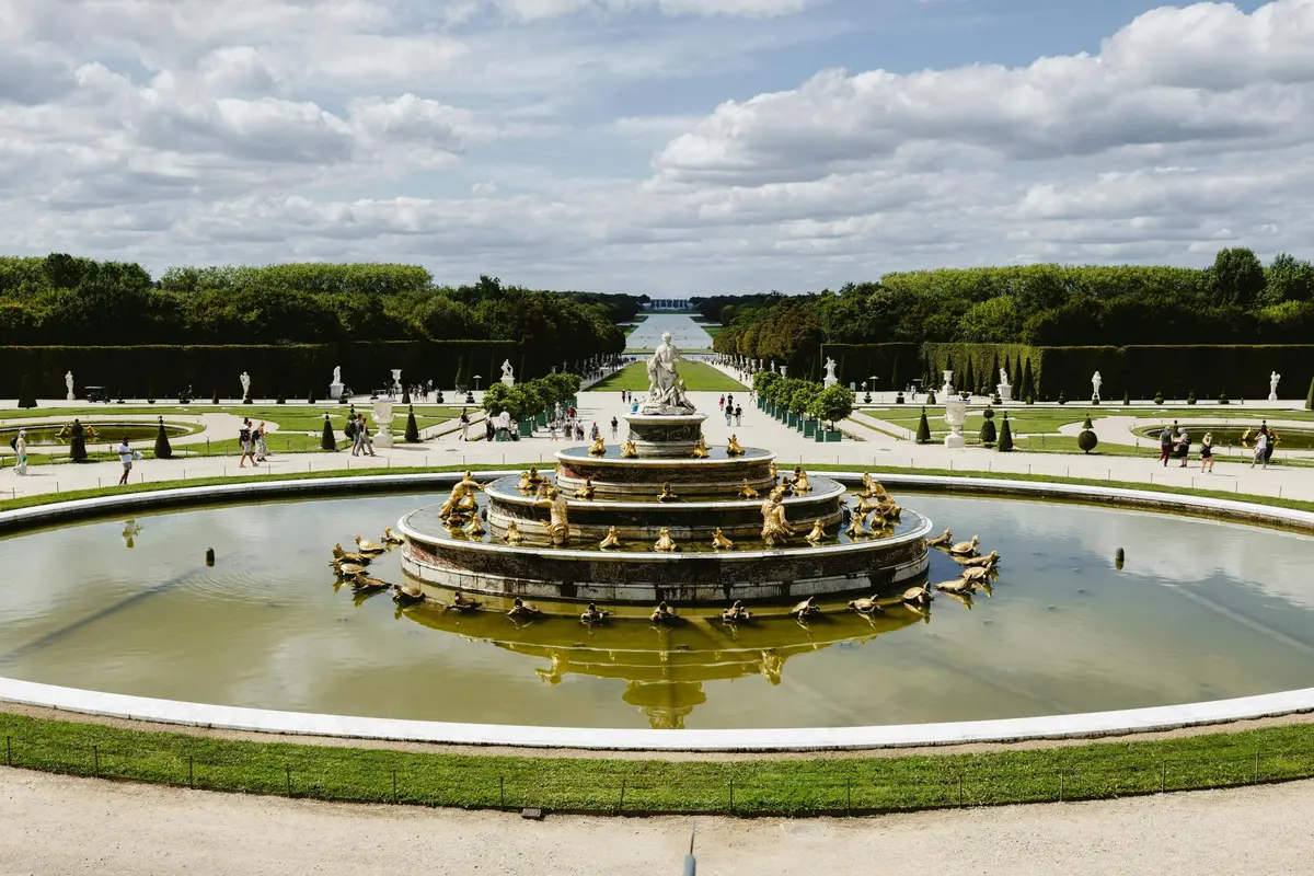 Une grande fontaine ornée de statues dorées dans un jardin à la française, entouré d'allées et de personnes, sous un ciel nuageux.