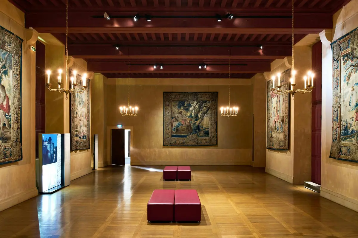 Chambre spacieuse avec sol en bois, bancs rouges, lustres suspendus et tapisseries décoratives sur les murs.