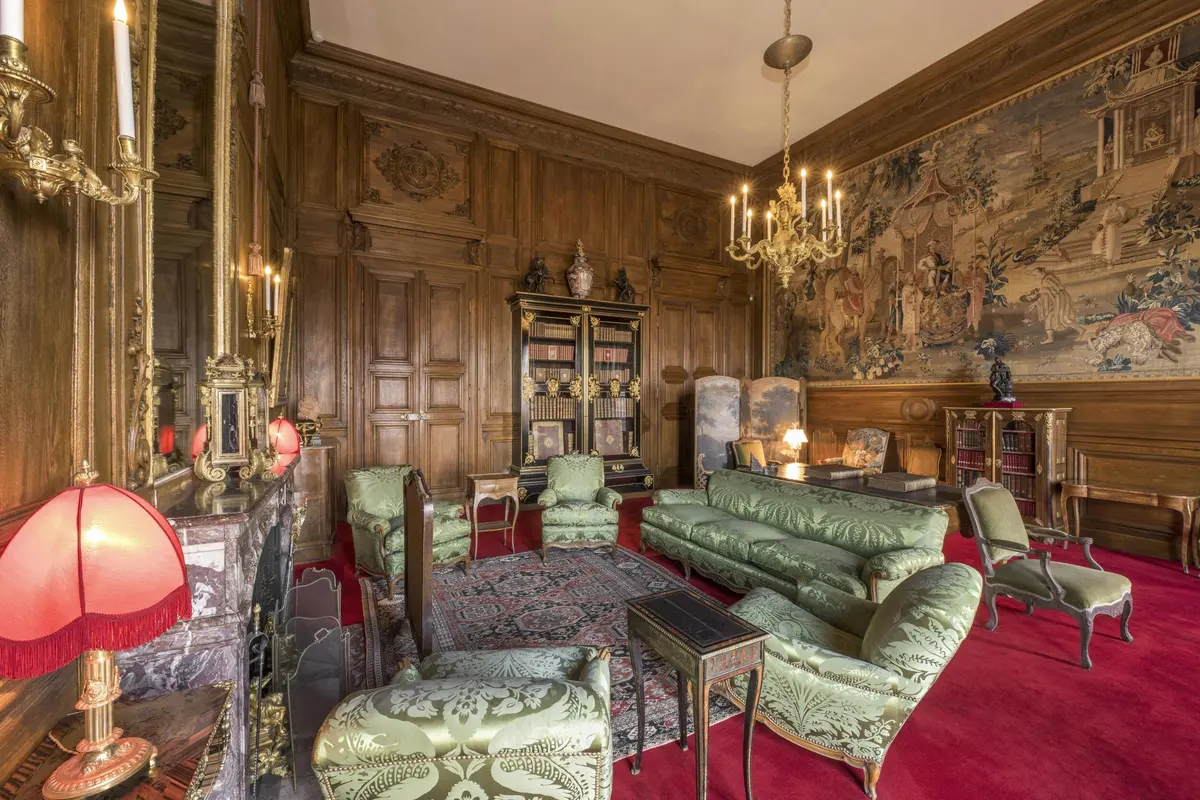Une chambre luxueuse avec des murs lambrissés, des meubles verts rembourrés, une grande bibliothèque, des lustres ornés et une tapisserie détaillée.